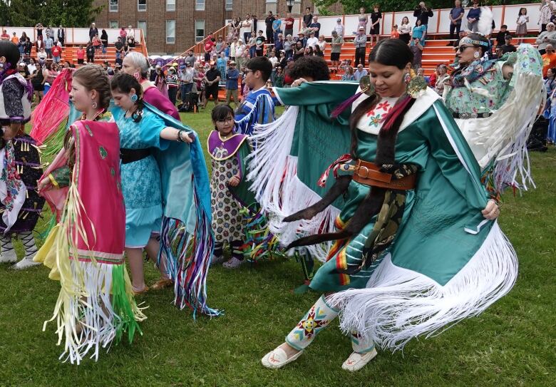 A group of people in regalia dance at a powow. There are stands in the background with people watching.