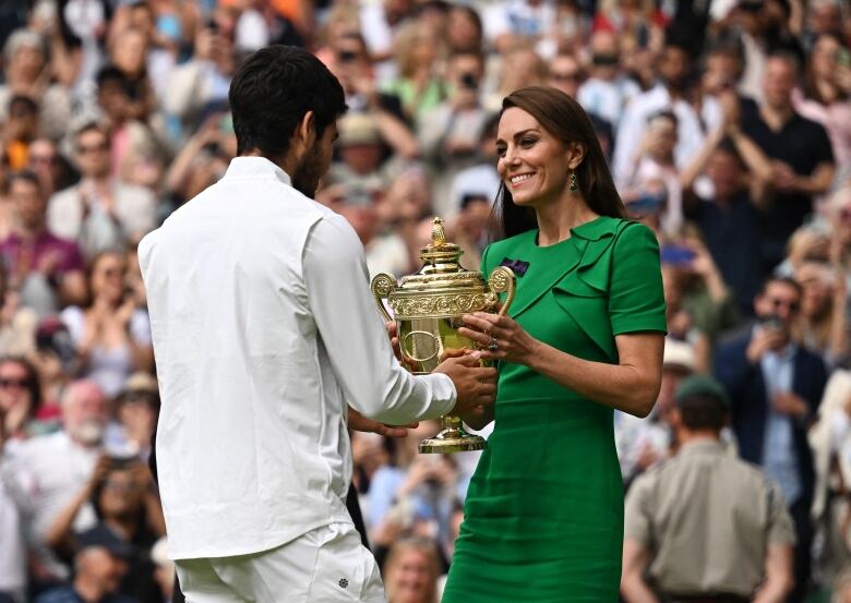 A person receives a trophy from another person while standing on a tennis court, with an audience in the background.