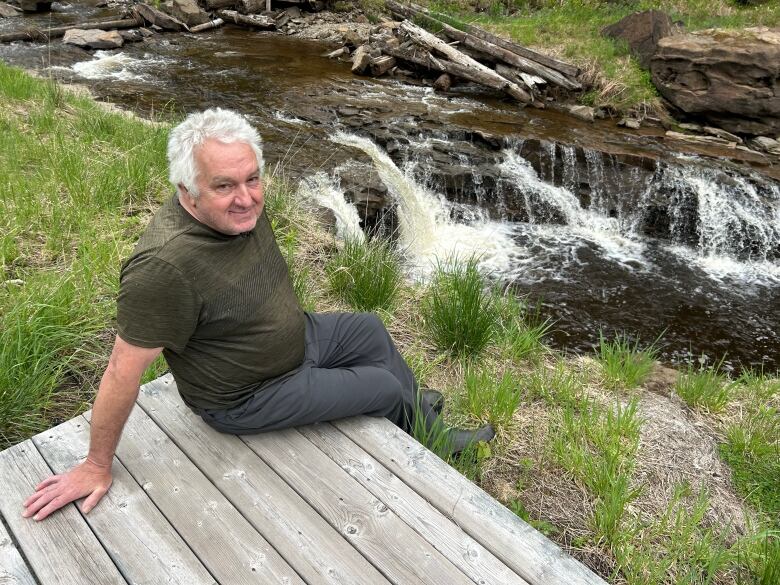 An older man with white hair sits on a wooden dock beside a rushing stream.