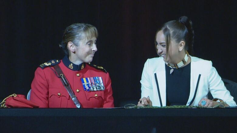 A woman in RCMP red serge and a woman in a white jacket over a black top sit at a table and smile.