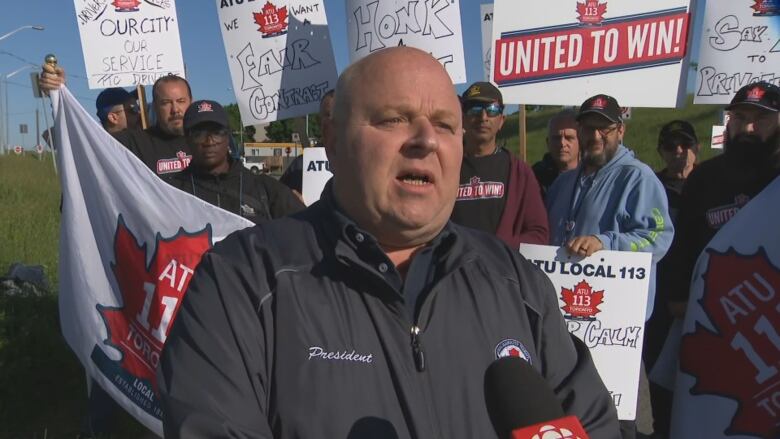 A bald middle aged man speaks into a CBC microphone. He is outside on a sunny day, in front of a group carrying picket signs