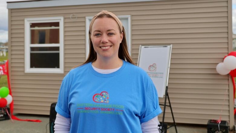 A woman in a blue t-shirt with a logo stands smiling in front of a small building