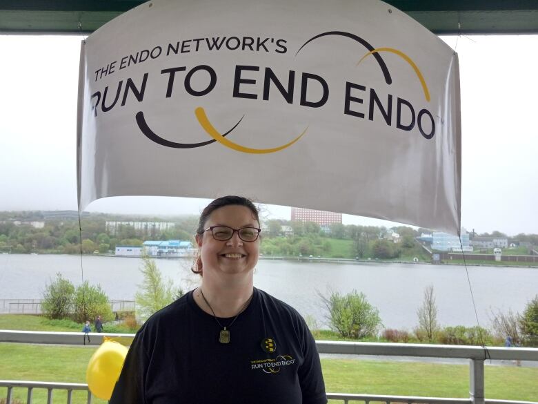 Woman with hair pulled into pony tail in black shirt stands before a sign that says Run to End Endo.