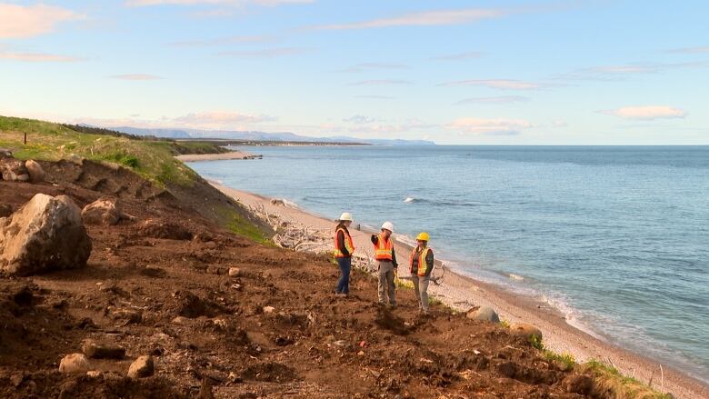 Three people wearing safety gear stand on a rocky embankment overlooking the ocean.