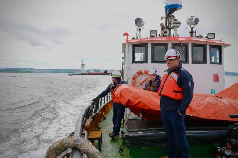 Three mariners stand on the deck of a tugboat with a barge and a tugboat on the water in the background. 