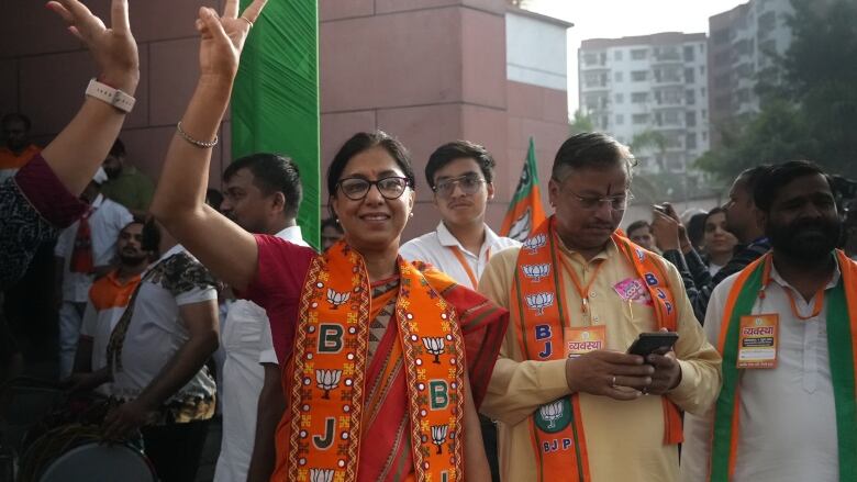 A dark-haired woman, wearing glasses and dressed in orange and red, gives the victory sign as a group of men stand nearby.