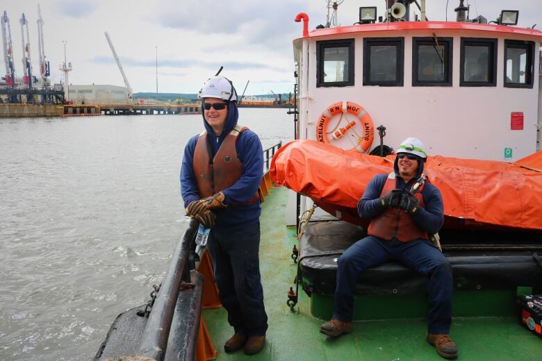 Two mariners in navy work clothes and dirty orange lifejackets laugh together on the deck of a tugboat. 