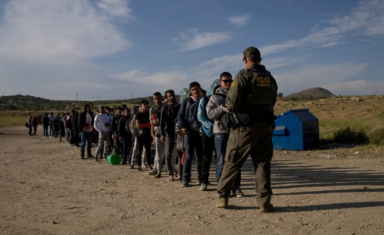 Dozens of men line up in front of a border patrol agent dressed in a green uniform.