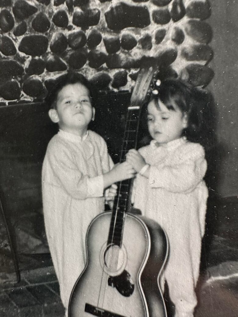 Two young children in their pyjamas hold onto an upright guitar. 