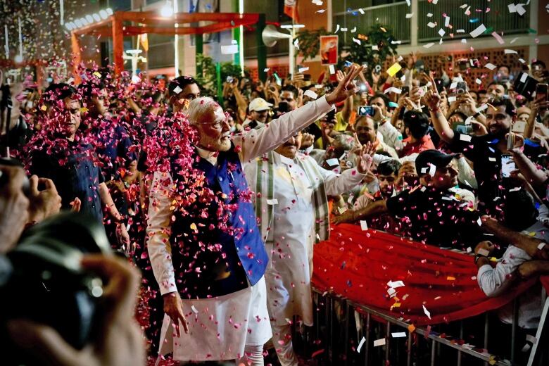 A white-haired man, wearing a white shirt and blue vest, waves to a loud crowd.