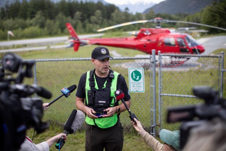 A man wearing a high-vis vest speaks to reporters, with a red helicopter behind him.