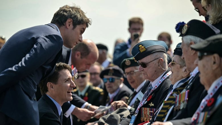 Elderly people in military uniforms are shown seated outdoors, in conversation with three cleanshaven men in suit and tie.