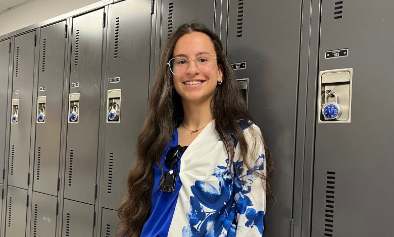 Girl in blue and white top, black pants and glasses stands near lockers.