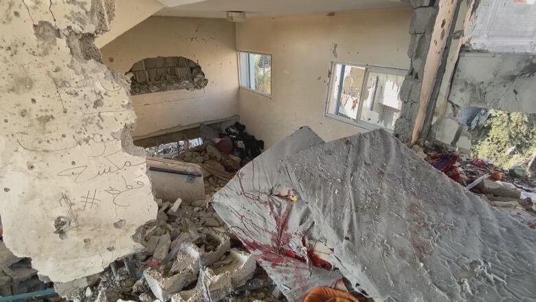 Two foam mattresses stacked on top of one another, the bottom one covered in blood, laying atop concrete debris near a wall blown out in a bombing. 