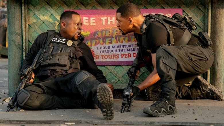 Two police officers wearing tactical gear crouch behind a short wall. 