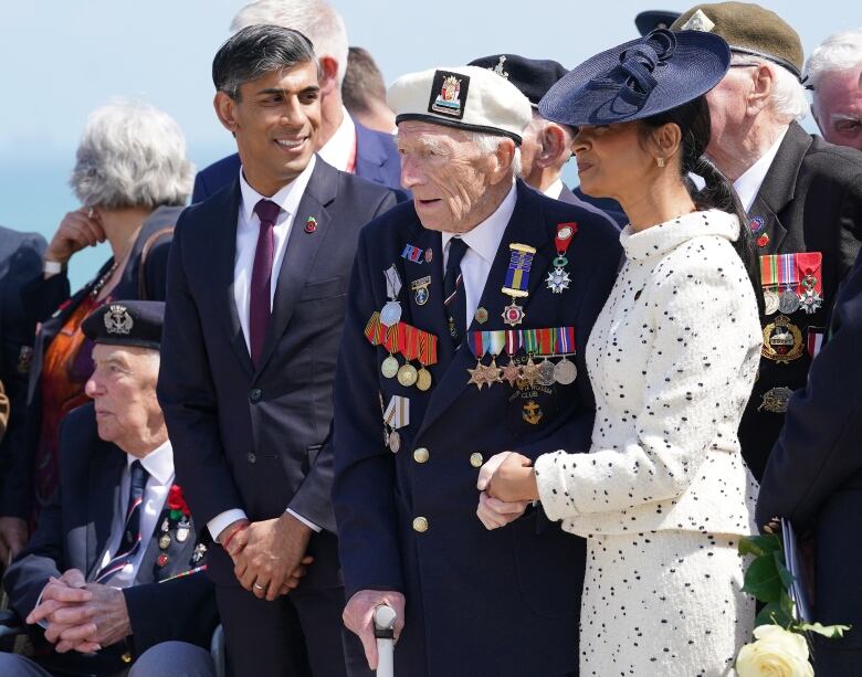 An elderly man in a military uniform stands with a younger man in a suit and tie on his right, and a woman in formal wear - dress and hat - to his left.