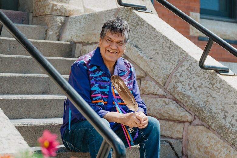 A person wearing a purple ribbon shirt holding an eagle feather sits on concrete stairs.