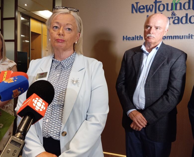 Woman with shoulder length blond hair an blue suit standing in front of a microphone.