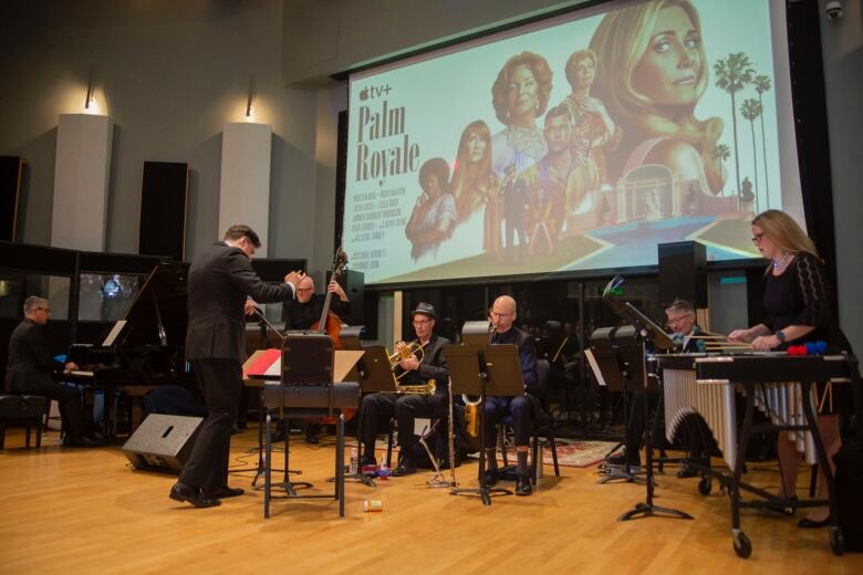 A conductor stands in front of musicians with a projection of the palm royale poster behind them