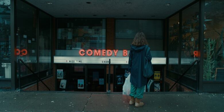 Actress Rachel Sennott stares at the sign for Comedy Bar in Toronto, while standing on the street holding bags.