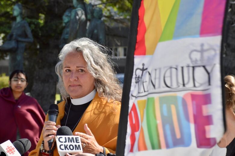 A woman wearing a white collar, black robe and a yellow jacket holds a microphone. A sign for Queen City Pride sits beside her. 