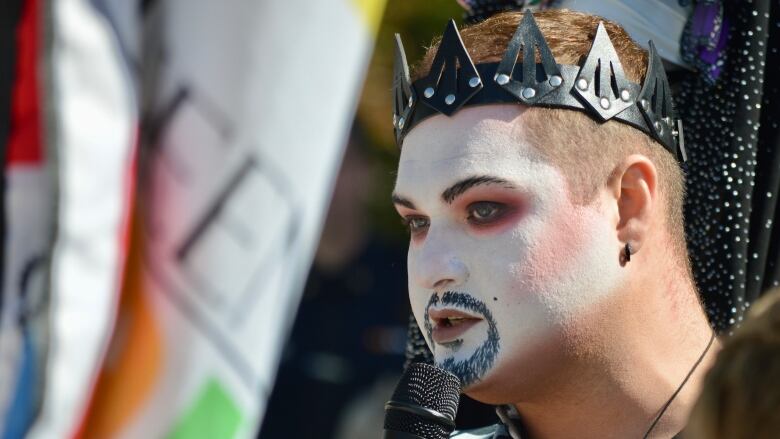 A person wearing make-up and a crown speaks into a microphone.