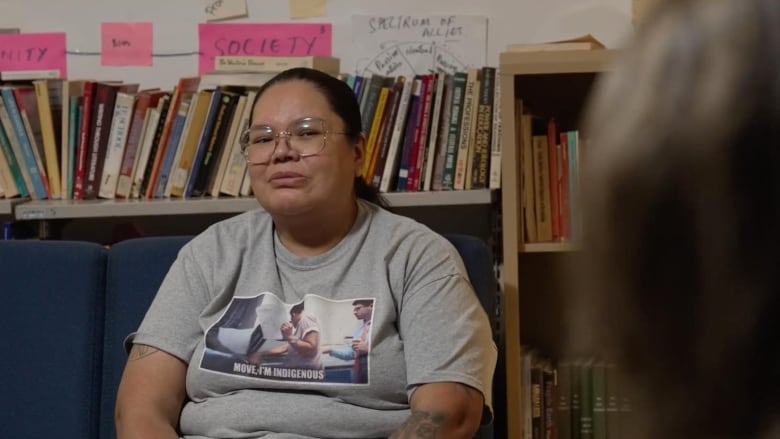 An Indigenous woman with glasses and a grey t-shirt stares at an interviewer off camera