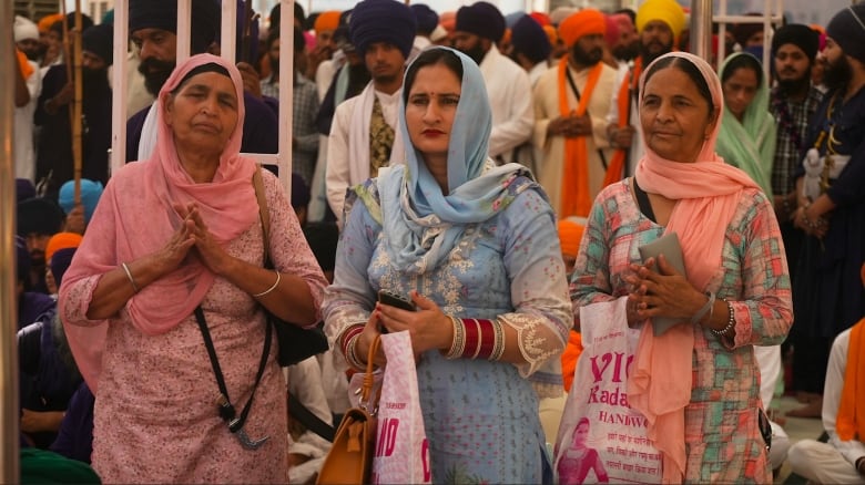 Three people with hands clasped stand in front of others taking part in a commemorative ceremoney.