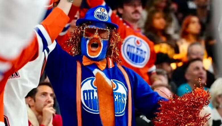 A man in blue-and-orange face paint, and a blue-and-orange Edmonton Oilers onesie, is standing up and cheering among the crowd at a hockey game.