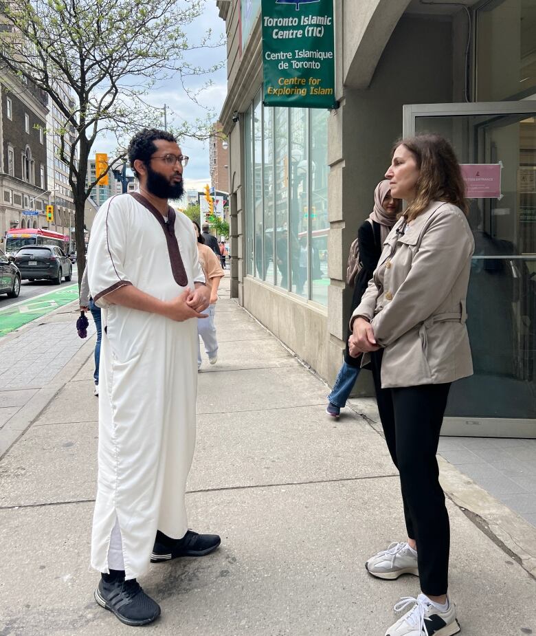 A man in a thawb stands on a city street, speaking with a reporter.