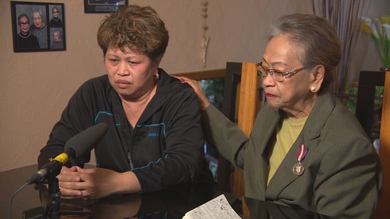 An older woman puts her hand on the shoulder of a woman who is looking down and appears sad.