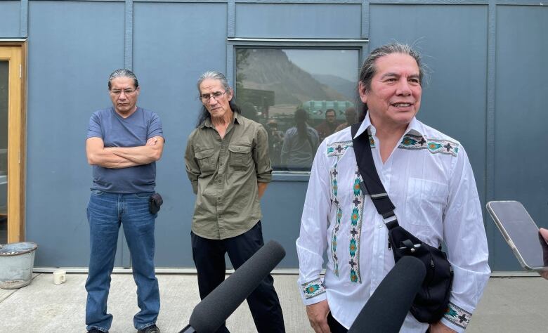 An Indigenous man speaks to reporters with mics, while two Indigenous men behind him look sullen at the ground.