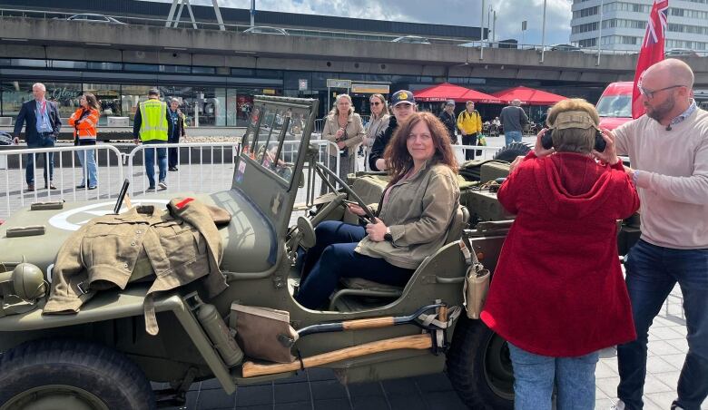 A woman sits behind the wheel in an army jeeps and looks at the camera as others look on.