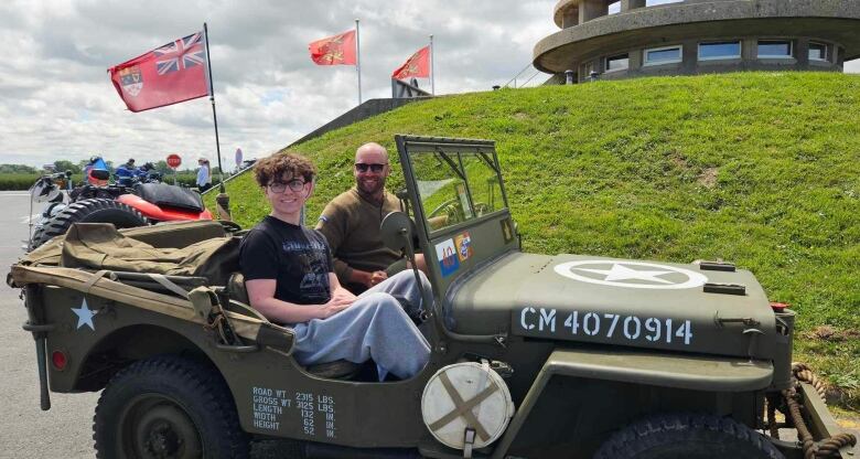 Two people sit in an army jeep with flags flying in the background. 