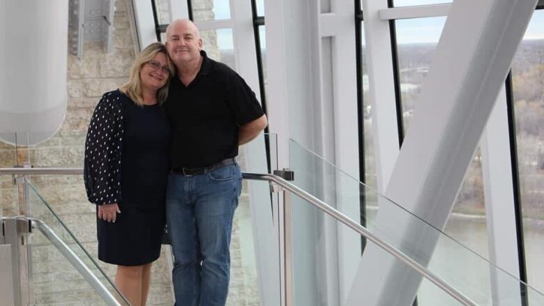 A man and a woman look to the camera. Windows to the right show a high-up view of a river and riverbank.