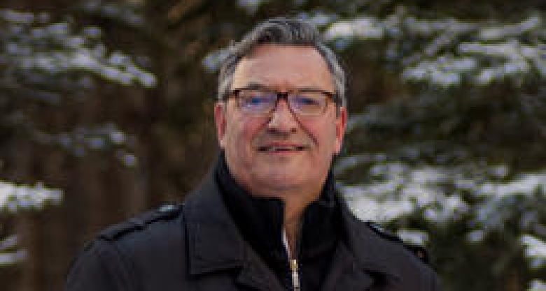 A man poses outside in front of snow-capped spruce trees.