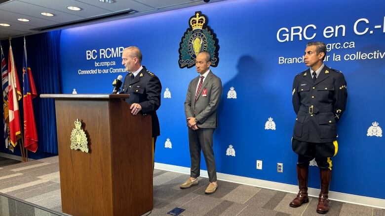 An RCMP officer stands at a podium with another officer and man in a grey suit behind him.