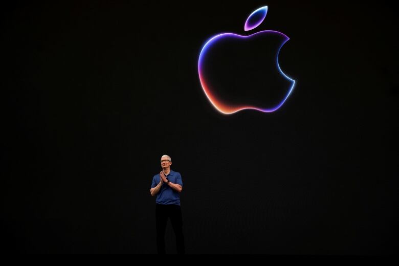 A man stands on a dark stage with a large multicoloured illustration of an apple glowing behind him.