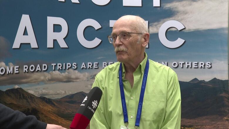 An older man stands in front of a banner with a CBC microphone held in front of him.