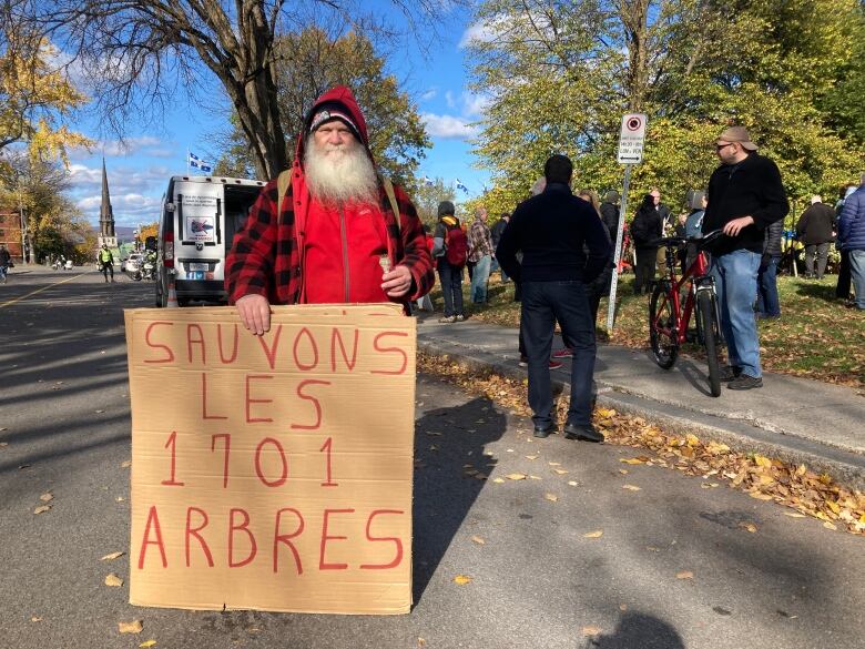 A man stands on a street with a cardboard sign that says 'save the 1701 trees'