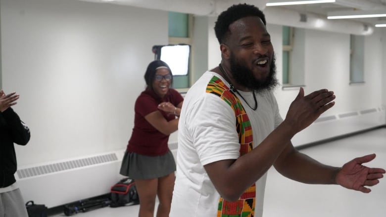 a bearded Black man wearing a white shirt mid-clap as his students dance in the background