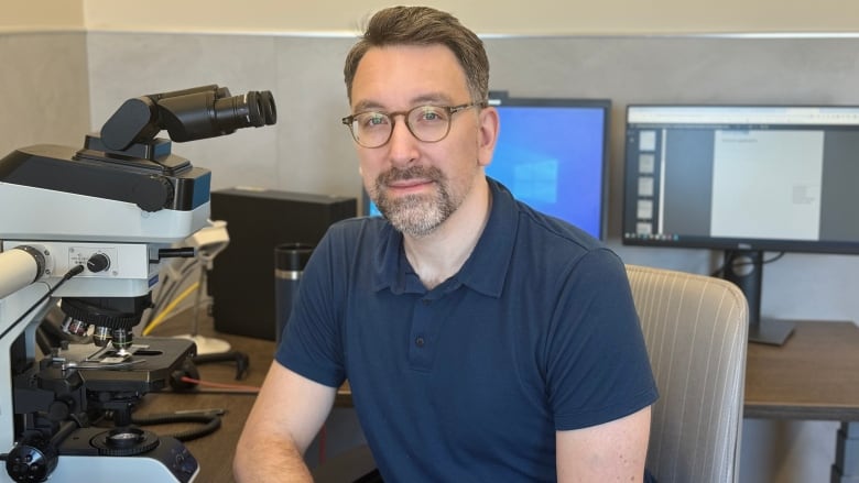 A man with a beard and glasses poses beside a microscope. He's wearing a short-sleeved navy shirt.