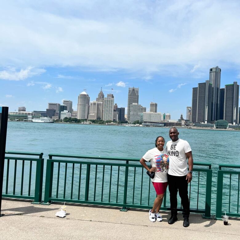 Man and woman stand in front of a metal fence. The Detroit River and Detroit skyline is behind them