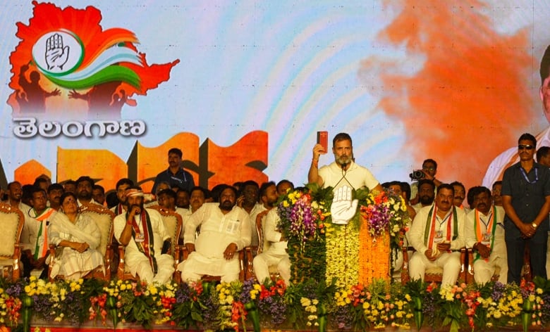 A man waves a red book at a podium. Around him are flowers and rows of people sitting behind him dressed in white.