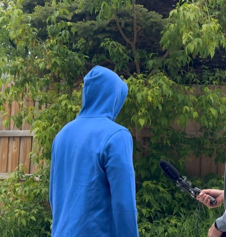 A person in a blue hoodie stands with their back to the camera near some leafy greenery. A person's hand holding a microphone peeks into the frame.