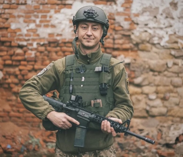 A portrait of a man in full military gear and a helmet. He holds an automatic weapon.