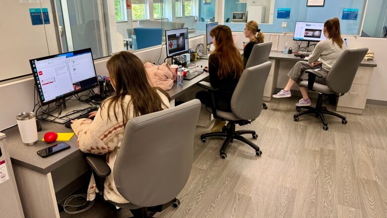 Four people sit at a large L-shaped desk, each of them working on a separate computer. There is a plexiglass divider between them and the outside hallway.