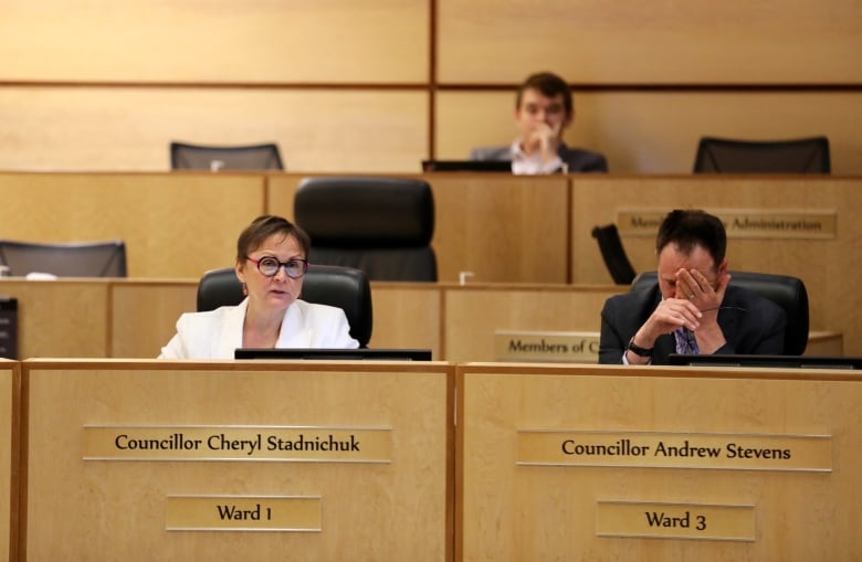 A woman in a white suit and a man in a black suit sit at a wooden desk. The man has his head in his hands. 