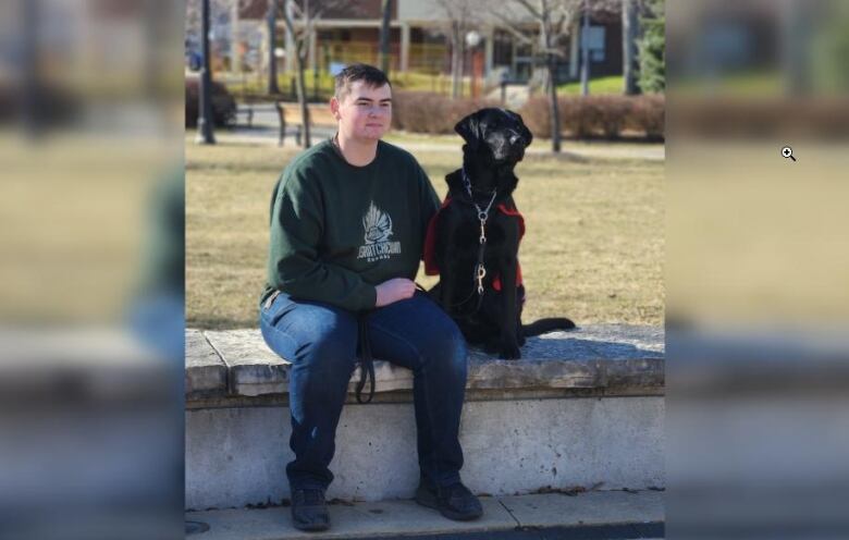 A teenager in a dark green sweater sits on a concrete ledge outdoors with a black dog in a red vest sitting beside him. 