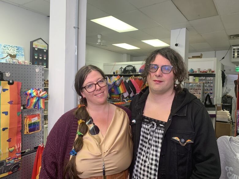 Two women pose for a photo surrounded by rainbow-coloured, Pride-themed merchandise. 